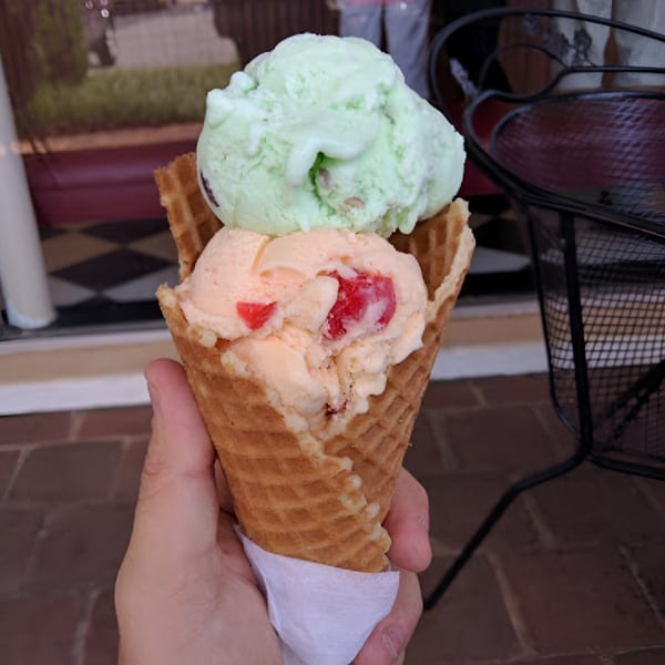 Hand holding a waffle cone with two scoops of ice cream, pistachio and cherry vanilla.