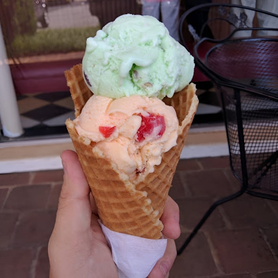 Hand holding a waffle cone with two scoops of ice cream, pistachio and cherry vanilla.
