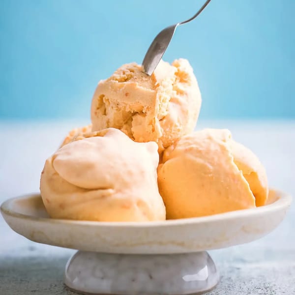 Three scoops of Mayberry's peach ice cream in a white bowl with a spoon, against a blue background.