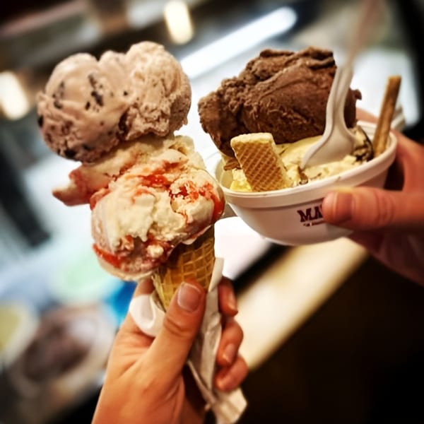 Two hands holding a Mayberry ice cream cone with strawberry and a cup with chocolate ice cream.