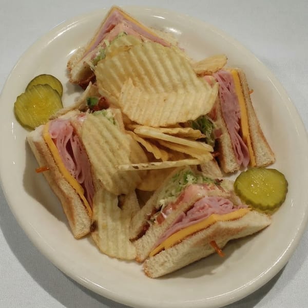 A club sandwich with ham, cheese, and lettuce, served with ruffled potato chips and pickles.