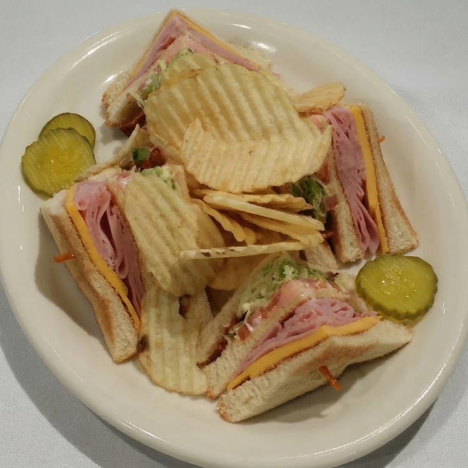 A club sandwich with ham, cheese, and lettuce, served with ruffled potato chips and pickles.